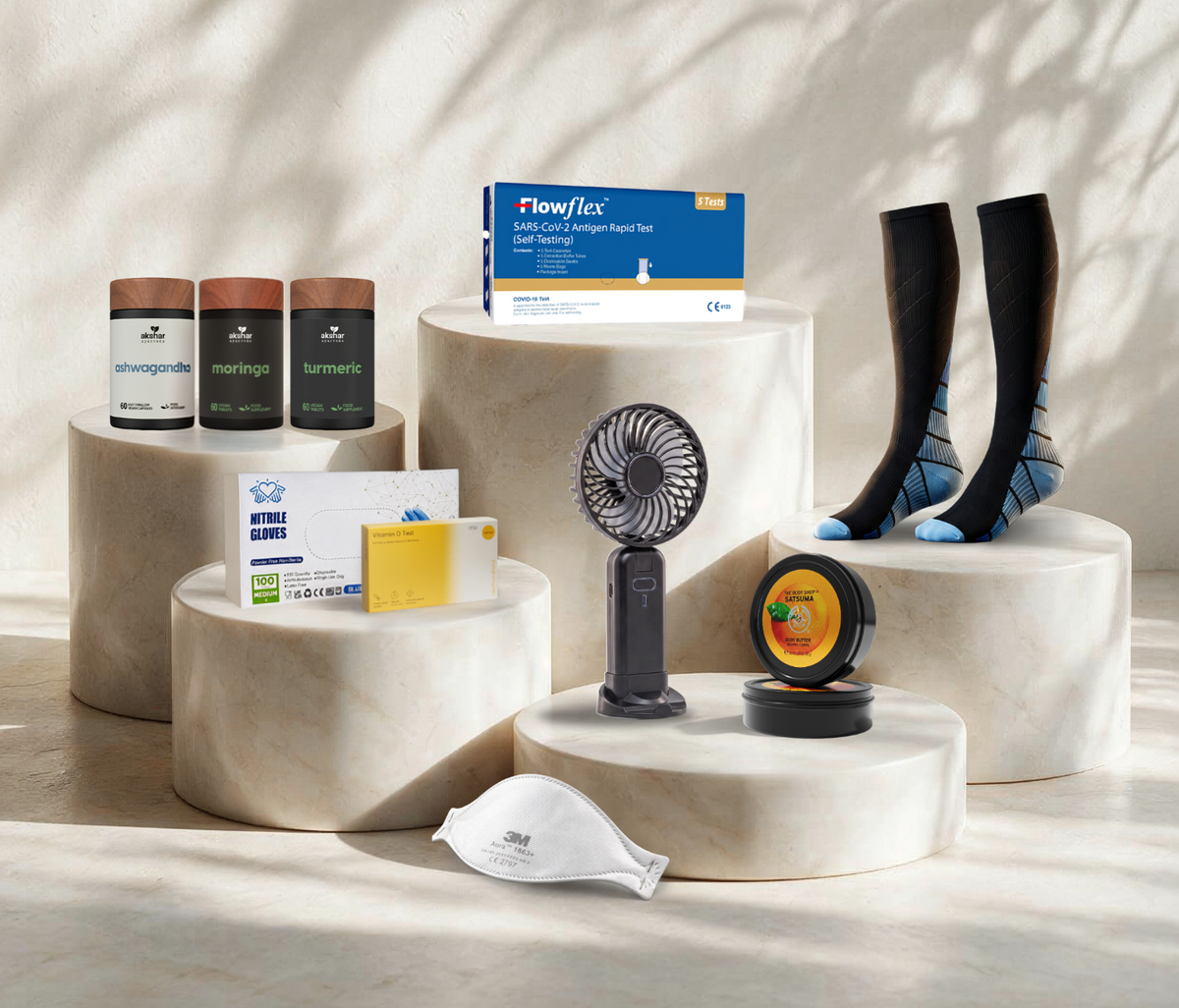 A curated product display featuring a variety of wellness and health items arranged on tiered cream stone pedestals under soft, dappled sunlight. The products include three supplement bottles, a Flowflex test box, a small desk fan, a face mask, and a pair of black compression socks with blue accents.